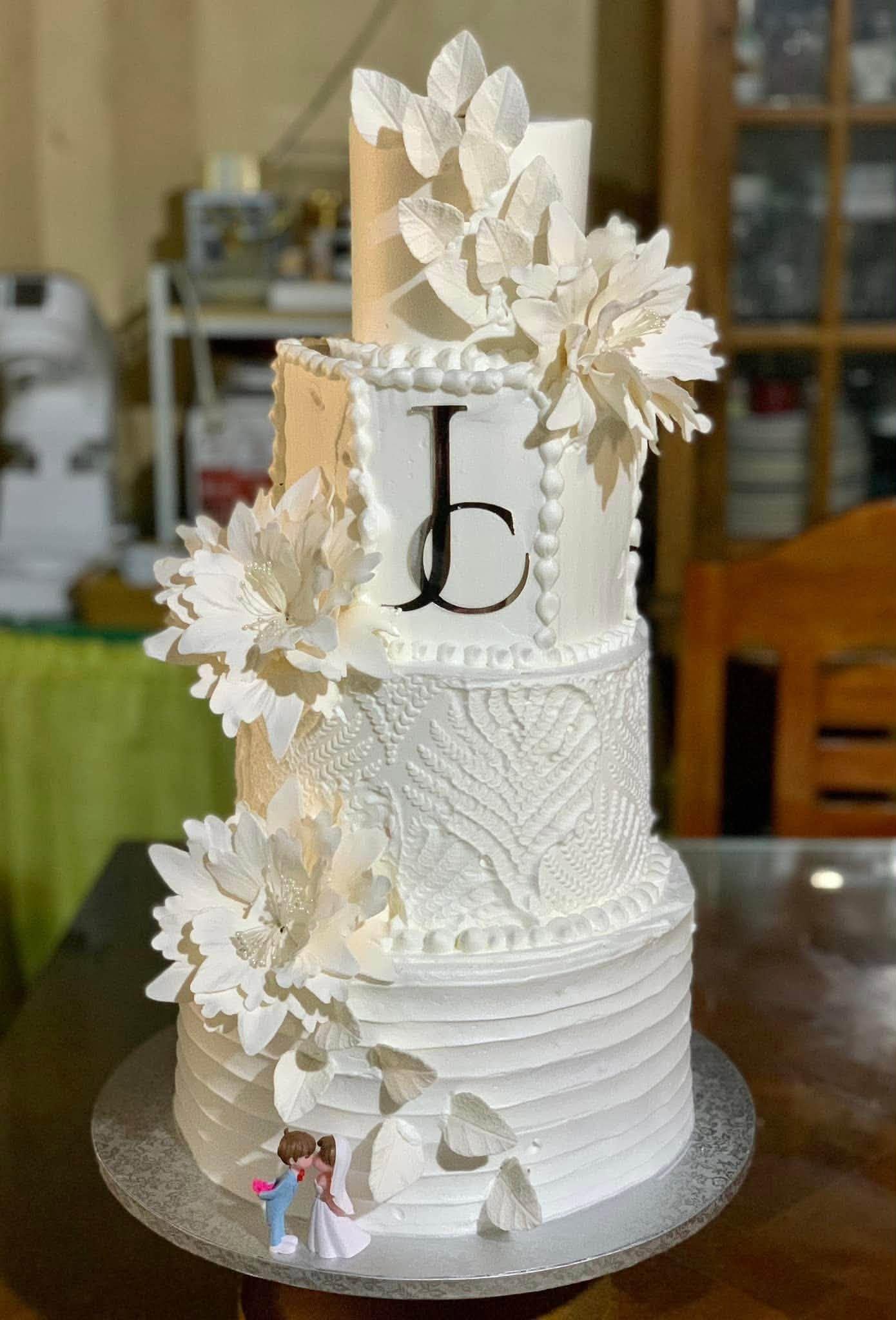 A tall four-tier white wedding cake decorated with textured patterns, large white sugar flowers, a gold monogram topper, and a small bride and groom figurine at the base.