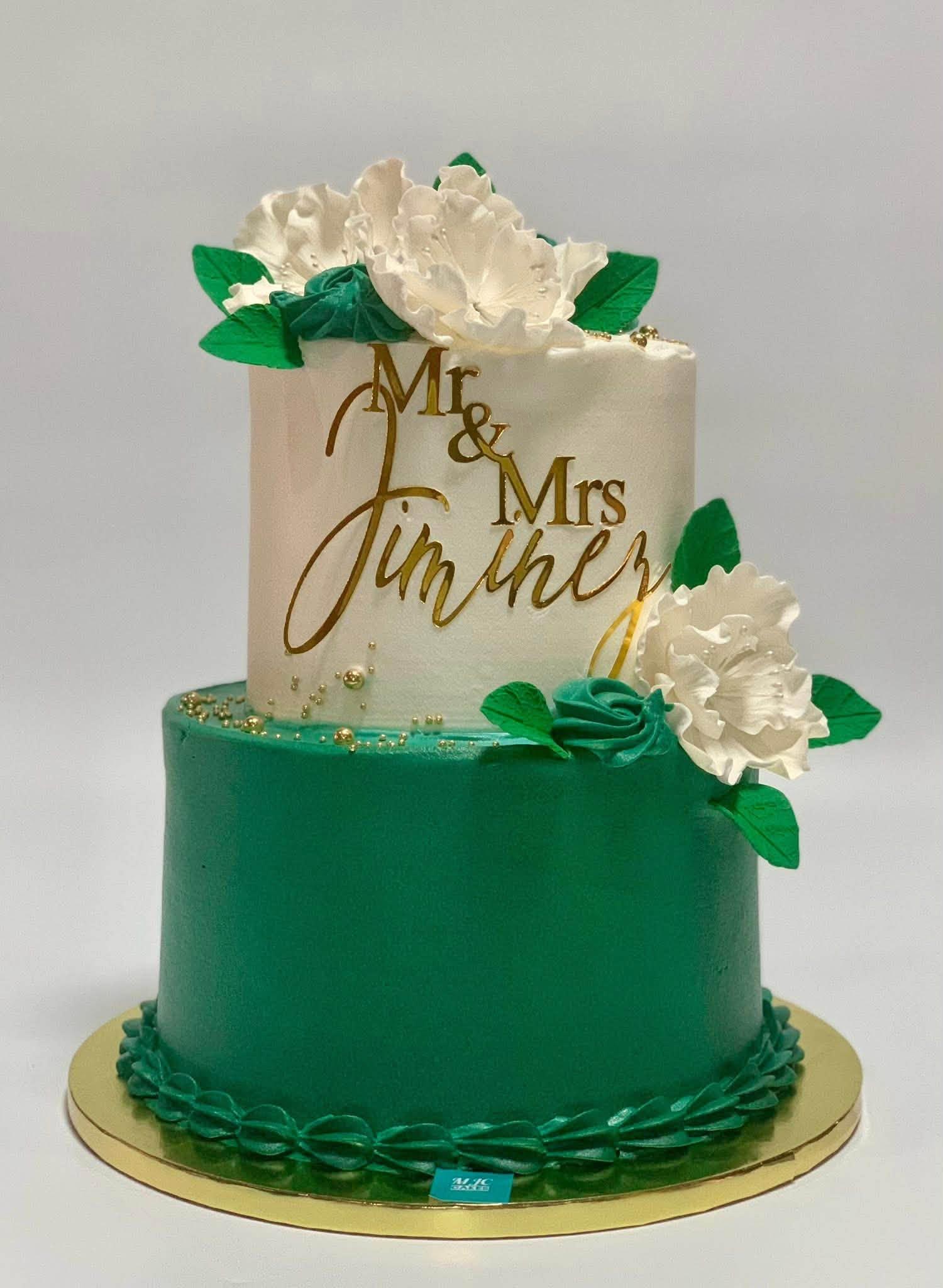 A two-tier wedding cake with a solid emerald green bottom tier and a white top tier decorated with large white sugar flowers, green leaves, gold pearls, and a gold acrylic topper that says Mr & Mrs Jimther.